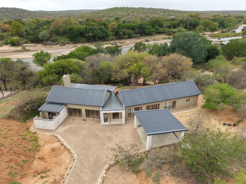 Aerial view of a modern house with a metal roof, carport, and paved driveway, situated in a natural bushveld setting near a river.