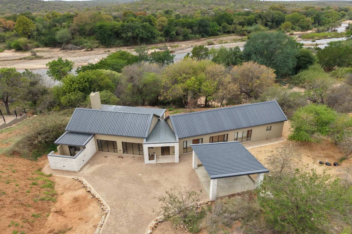 Aerial view of a modern house with a metal roof, carport, and paved driveway, situated in a natural bushveld setting near a river.
