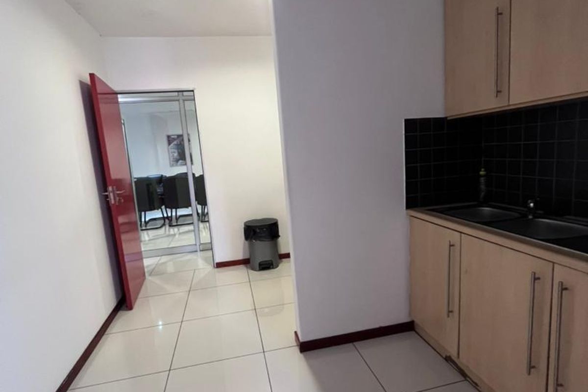 A compact kitchenette featuring light wood cabinetry, black tiled splashback, and tiled flooring, with an adjacent doorway leading to an office space.