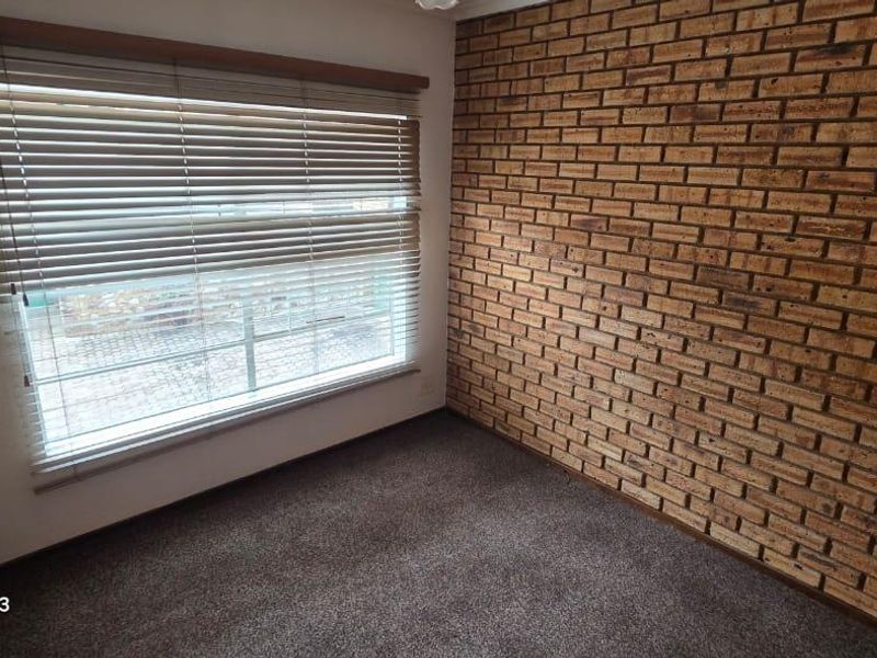 A vacant bedroom featuring a distinctive exposed brick feature wall, carpeted flooring, and fitted venetian blinds over the window.