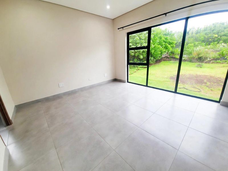 A bright, modern, and empty bedroom featuring large-format floor tiles, recessed lighting, and expansive black aluminium sliding doors opening to a garden view.