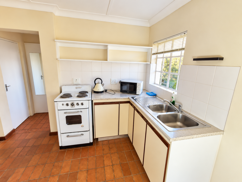 A functional kitchen featuring a freestanding stove, double bowl stainless steel sink, tiled splashbacks, and ample built-in cabinetry with terracotta floor tiles.