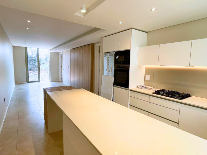 Modern open-plan kitchen and living area featuring sleek white cabinetry, integrated appliances, stone countertops, and neutral tiled flooring in a bright, contemporary space.