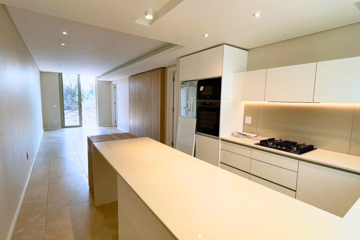 Modern open-plan kitchen and living area featuring sleek white cabinetry, integrated appliances, stone countertops, and neutral tiled flooring in a bright, contemporary space.
