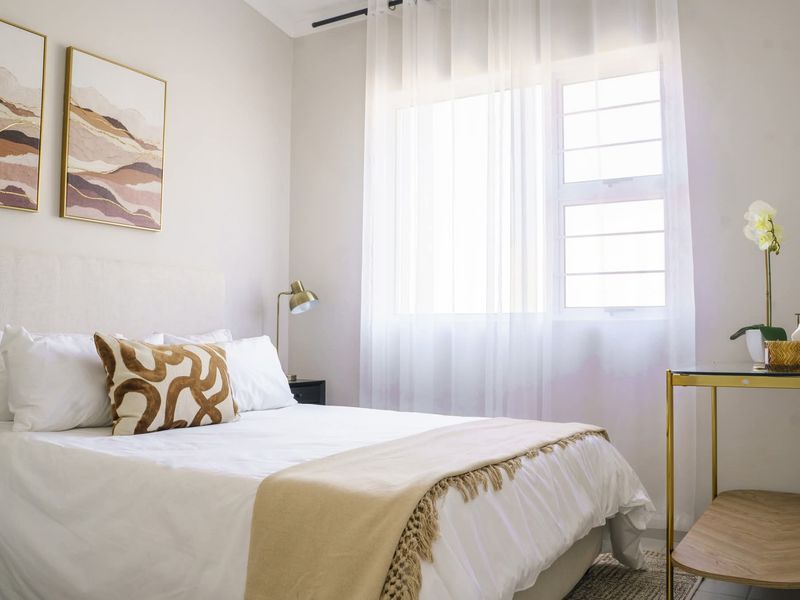 A well-lit, modern bedroom featuring neutral tones, a large window with sheer curtains, and clean, well-maintained finishes.