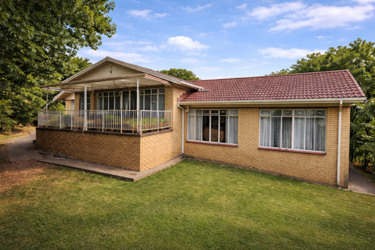 A rear view of a single-story brick house featuring a covered patio area, large windows, and a surrounding lawn.