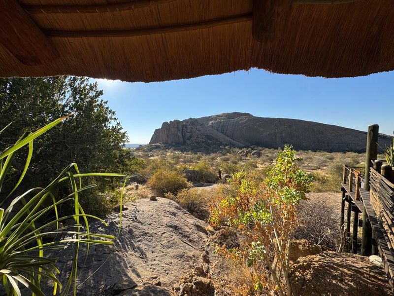 A scenic view of the rocky landscape and natural terrain, framed by a traditional lodge thatch roof and timber deck structure.