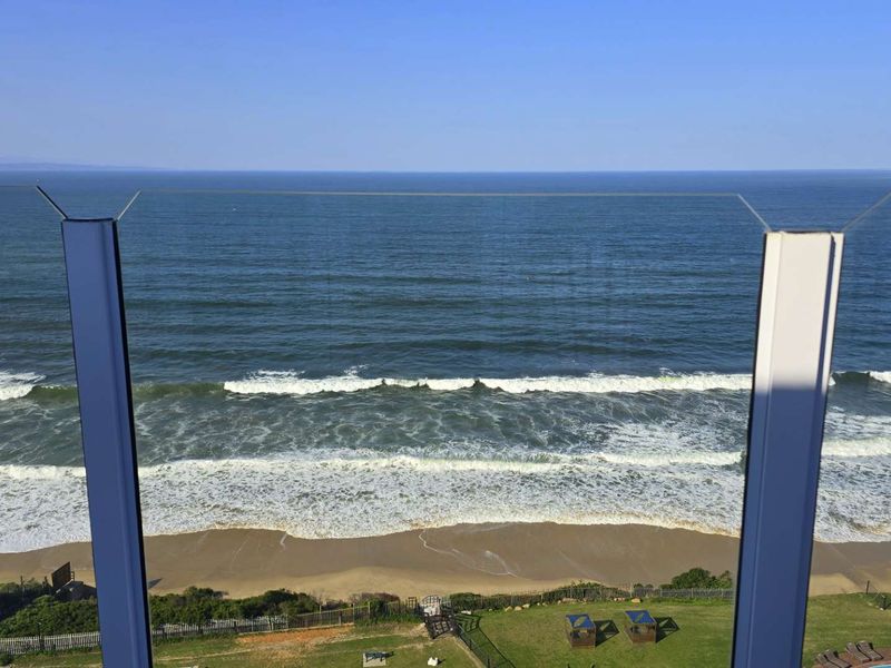 A clear, elevated ocean view captured from the penthouse balcony, featuring a modern glass balustrade overlooking the beach and coastline.