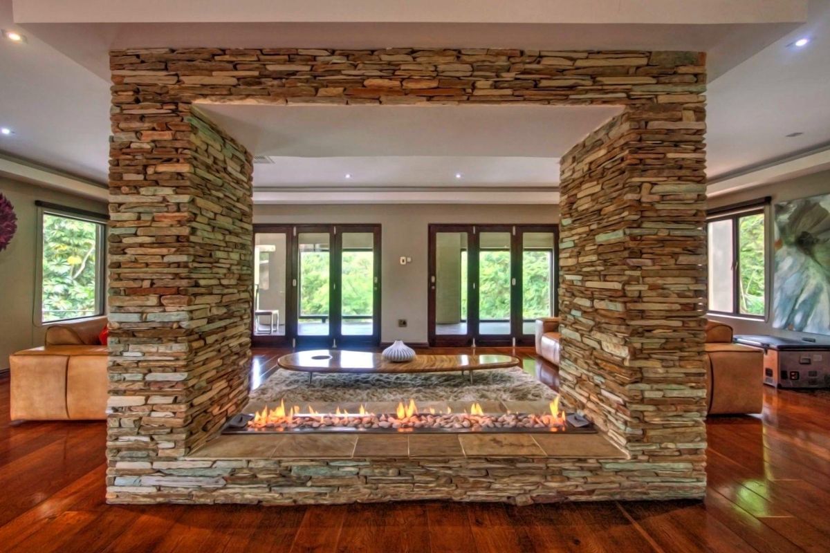 Spacious living area featuring a prominent central stone-clad fireplace, hardwood flooring, and large glass doors providing ample natural light and garden views.