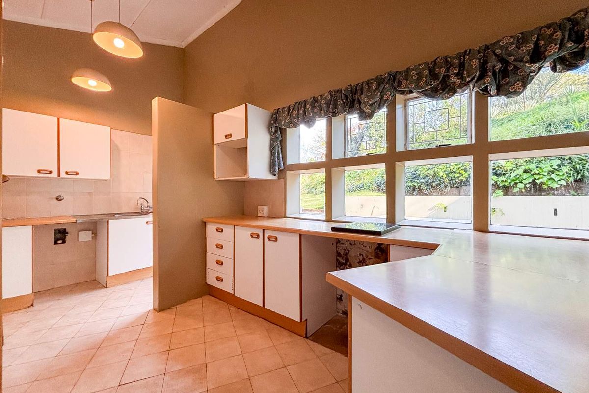 A dated kitchen featuring white cabinetry, tiled flooring, and large windows overlooking the garden, offering significant potential for a modern renovation.