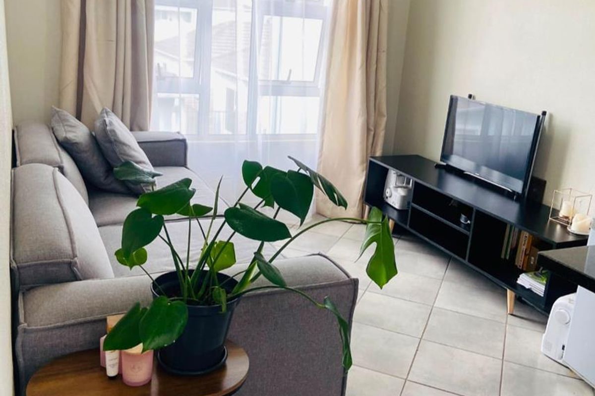 A compact living room featuring neutral tiled flooring, cove cornicing, and a large window allowing natural light into the space.
