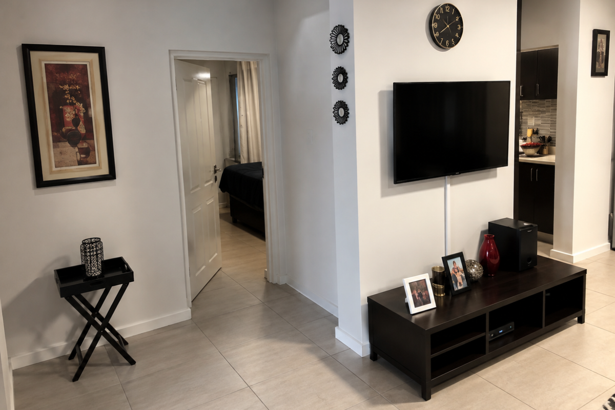 A well-maintained living area featuring neutral tiled flooring, a wall-mounted television, and direct access to an adjacent bedroom.