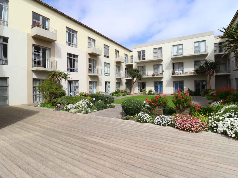 A well-maintained apartment complex courtyard featuring a central landscaped garden, paved pathways, and timber decking, surrounded by multi-story residential units with private balconies.
