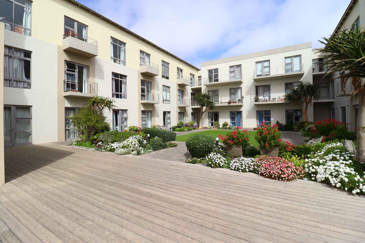 A well-maintained apartment complex courtyard featuring a central landscaped garden, paved pathways, and timber decking, surrounded by multi-story residential units with private balconies.