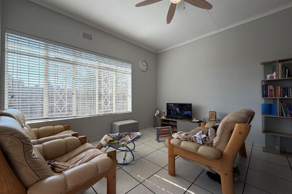 A living room featuring tiled flooring, a ceiling fan, and large windows with venetian blinds, providing a functional and well-lit space.
