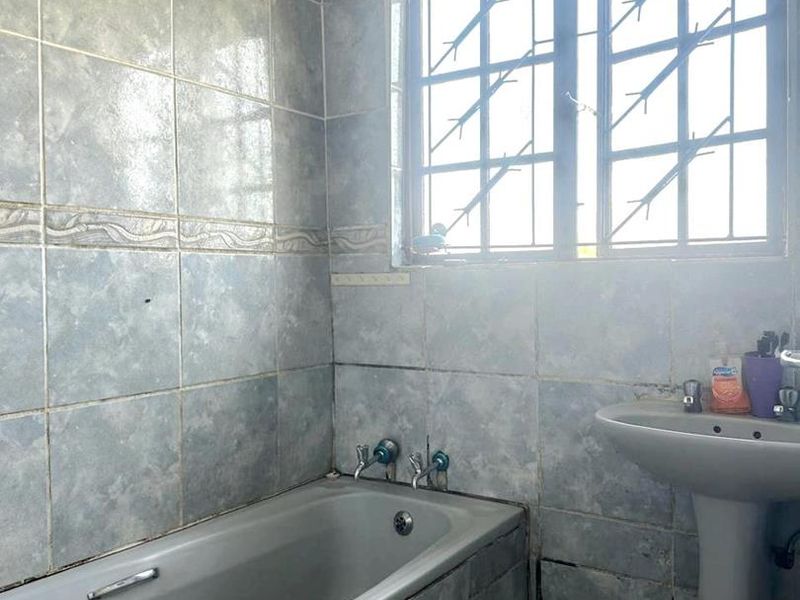A dated bathroom featuring a built-in bathtub, pedestal basin, and full-height wall tiling with security bars on the window.