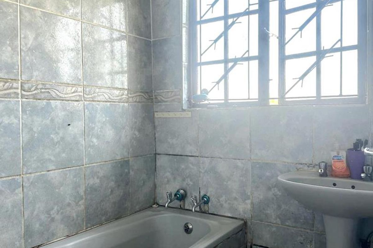 A dated bathroom featuring a built-in bathtub, pedestal basin, and full-height wall tiling with security bars on the window.