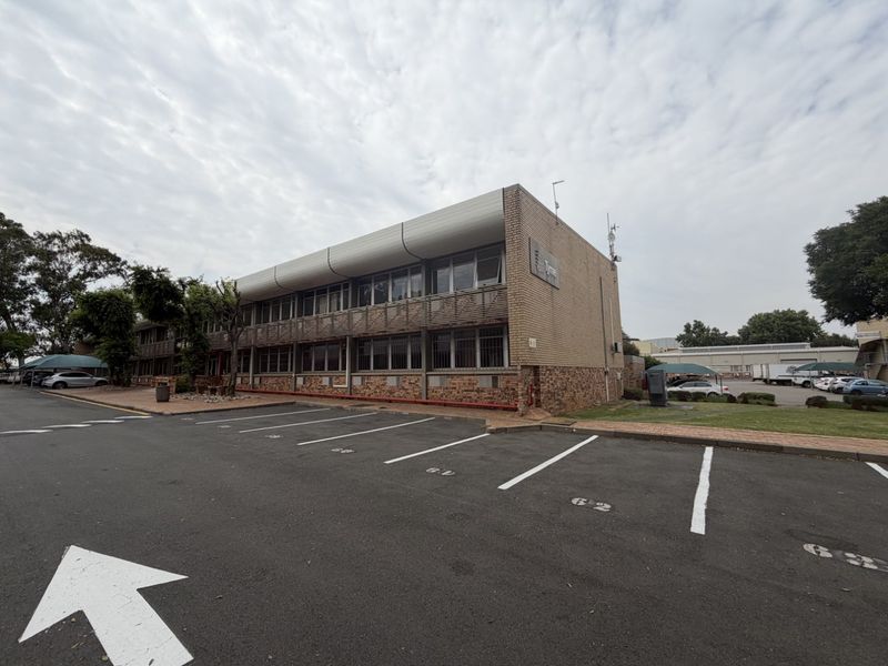 Exterior view of a multi-story industrial office building featuring brick facade, metal-framed windows with security bars, and an adjacent asphalt parking area.