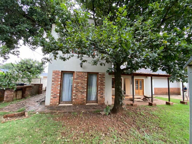 Rear view of the cottage featuring a covered patio area, brick paving, and an outdoor built-in braai, set within a garden with mature trees.
