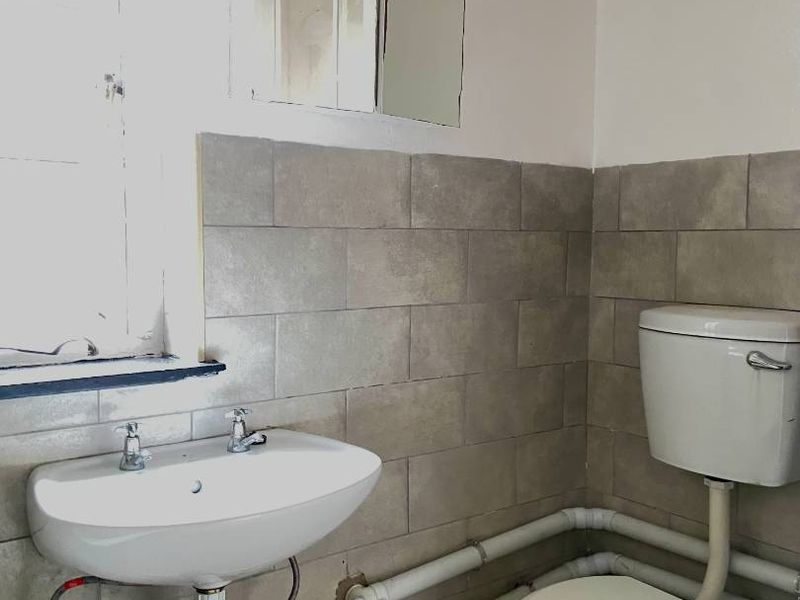 A functional bathroom featuring a wall-mounted basin, toilet, and tiled splashback, currently showing dated finishes and exposed plumbing.