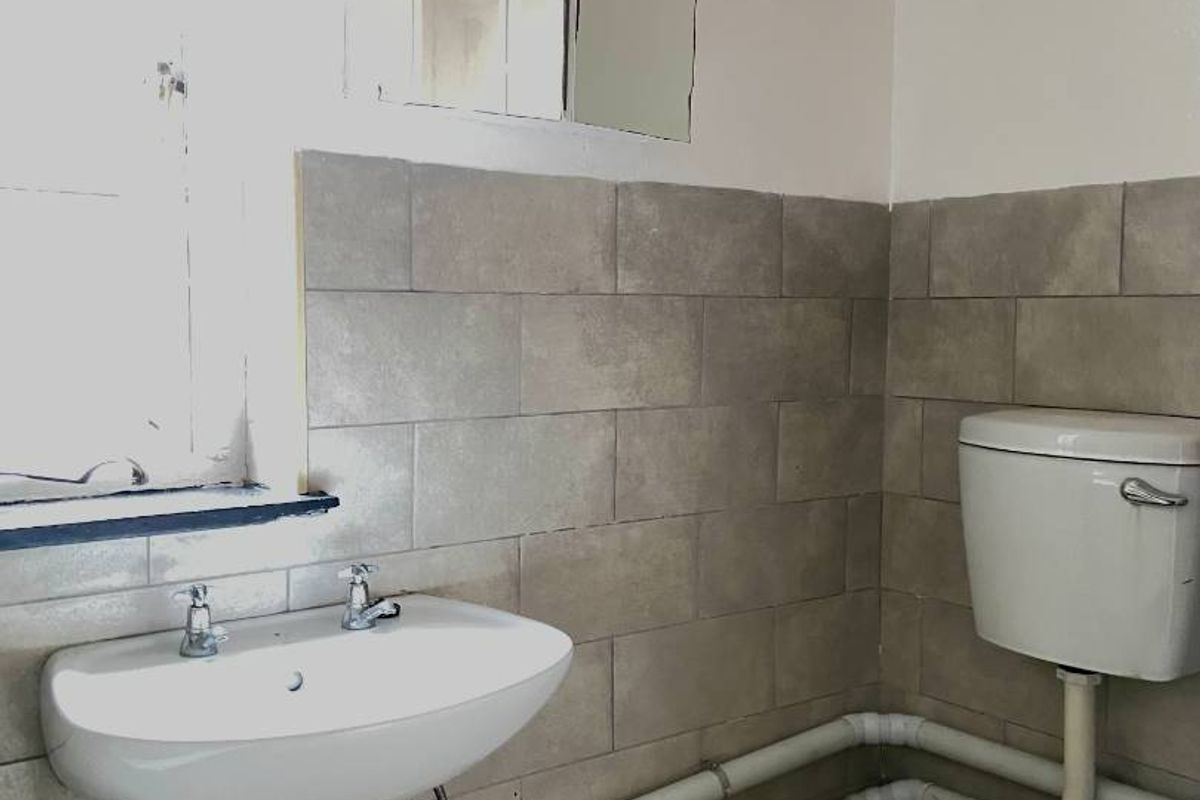 A functional bathroom featuring a wall-mounted basin, toilet, and tiled splashback, currently showing dated finishes and exposed plumbing.