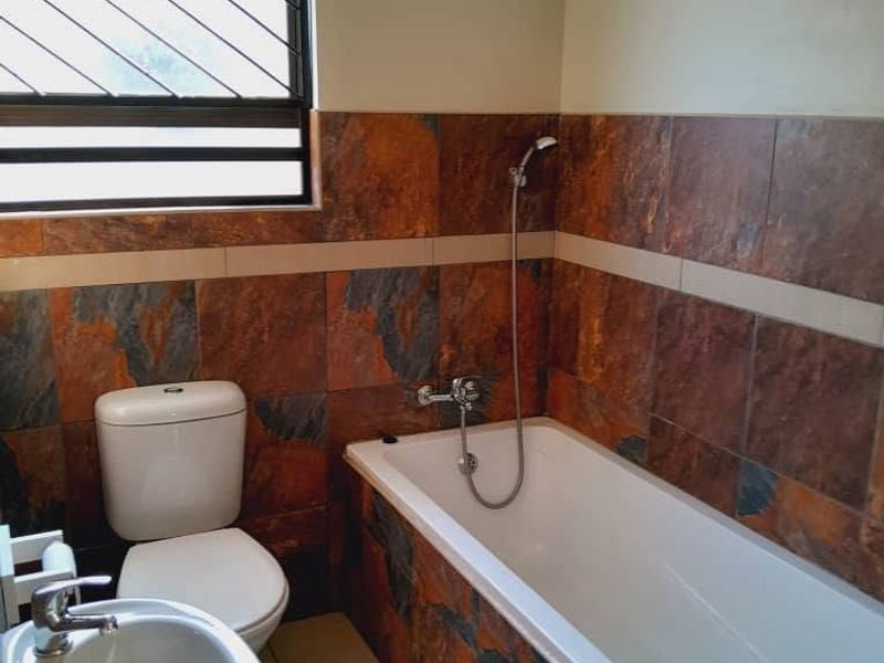 A functional bathroom featuring a bathtub with handheld shower, pedestal basin, and toilet, finished with distinctive dark stone-look wall tiles.