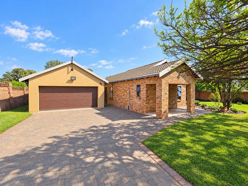 Front view of a well-maintained brick house featuring a paved driveway, an attached garage, and a covered entrance porch set within a landscaped garden.
