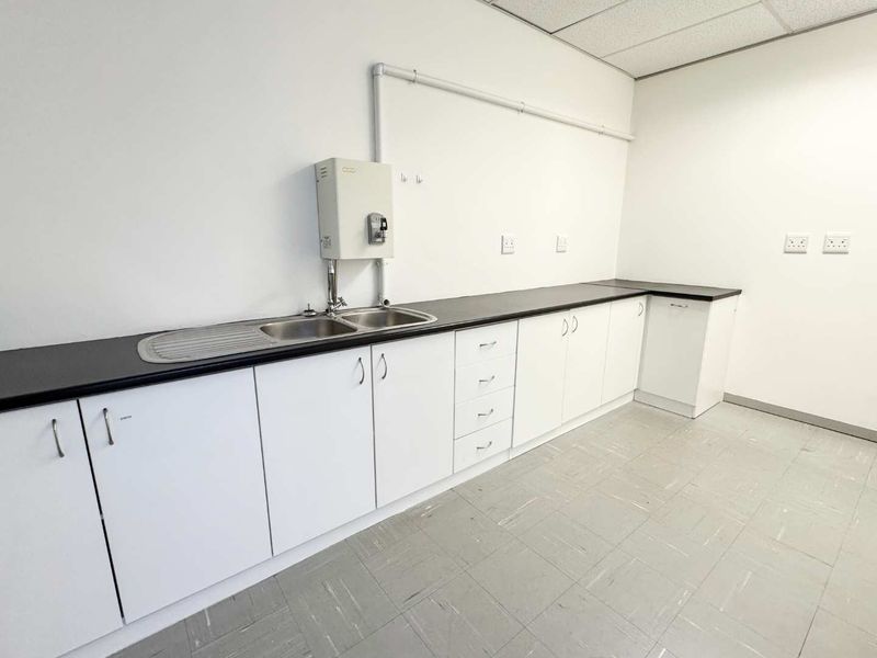 A clean, functional kitchenette area featuring white cabinetry, dark laminate countertops, a double sink, and durable tiled flooring.