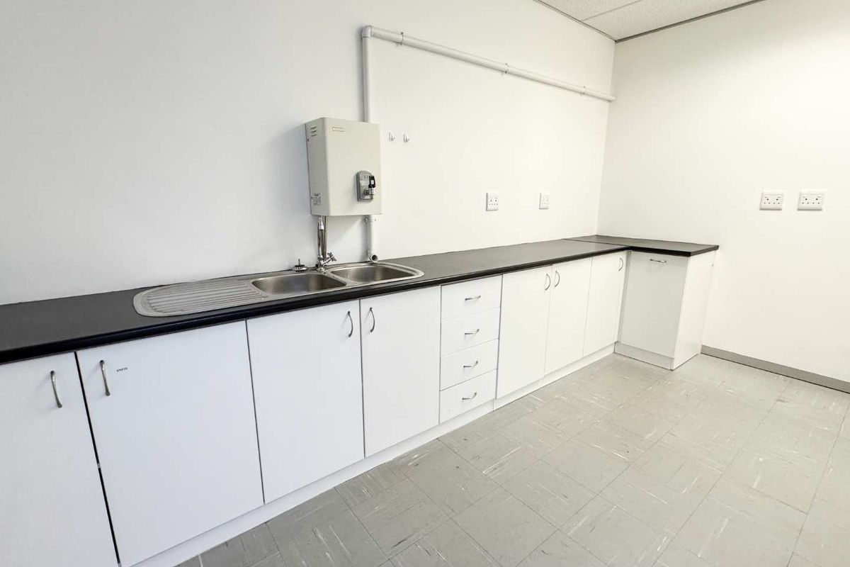 A clean, functional kitchenette area featuring white cabinetry, dark laminate countertops, a double sink, and durable tiled flooring.