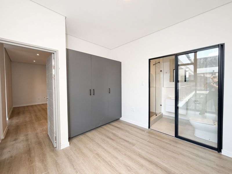 Modern bedroom featuring light laminate flooring, built-in grey cupboards, and a glass-enclosed en-suite bathroom, offering a clean and contemporary living space.
