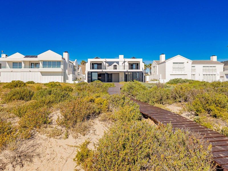 Rear view of a modern coastal residence featuring a wooden boardwalk leading through dunes to the property with balconies and large sliding glass doors.