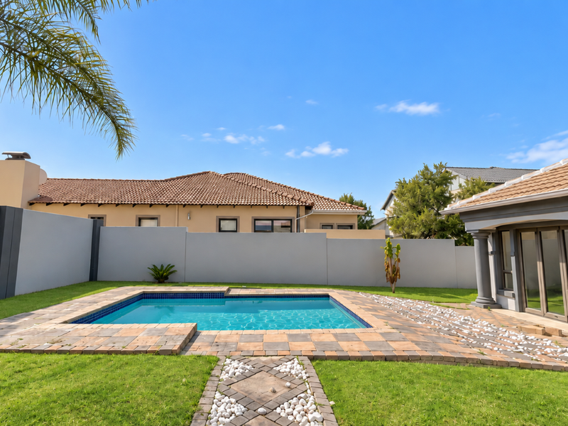 A well-maintained outdoor pool area featuring a paved surround, manicured lawn, and a covered patio structure with decorative columns.
