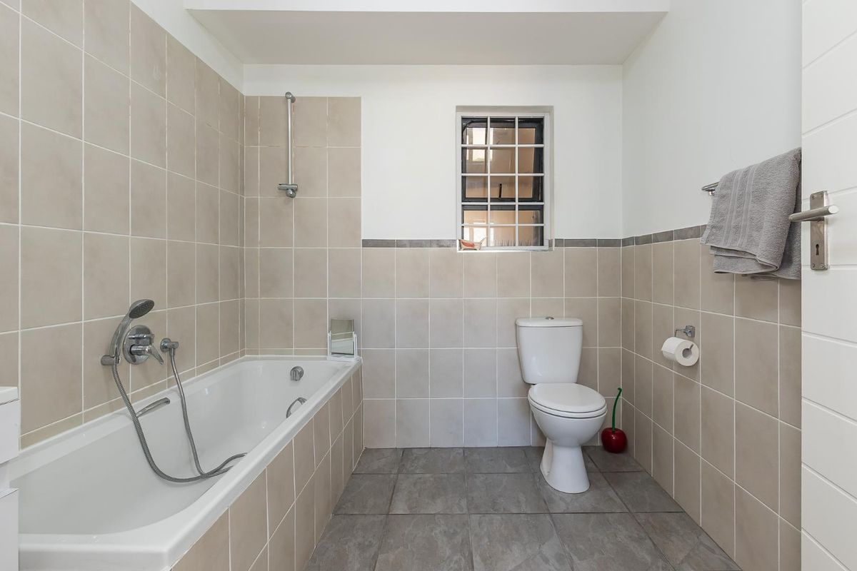 A functional bathroom featuring a built-in bathtub with shower attachment, a standard toilet, and neutral wall tiling with a window for natural light.