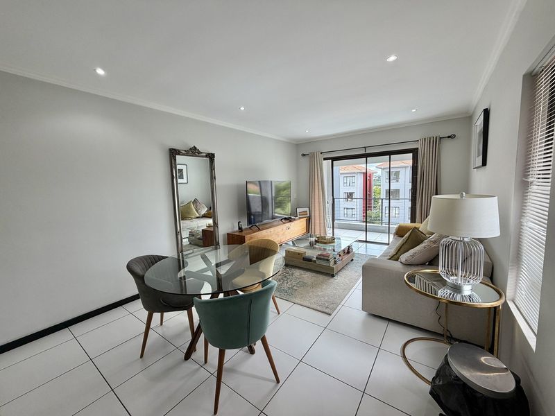 A bright open-plan living and dining area featuring tiled flooring, recessed downlighting, and a sliding glass door leading to a private balcony.