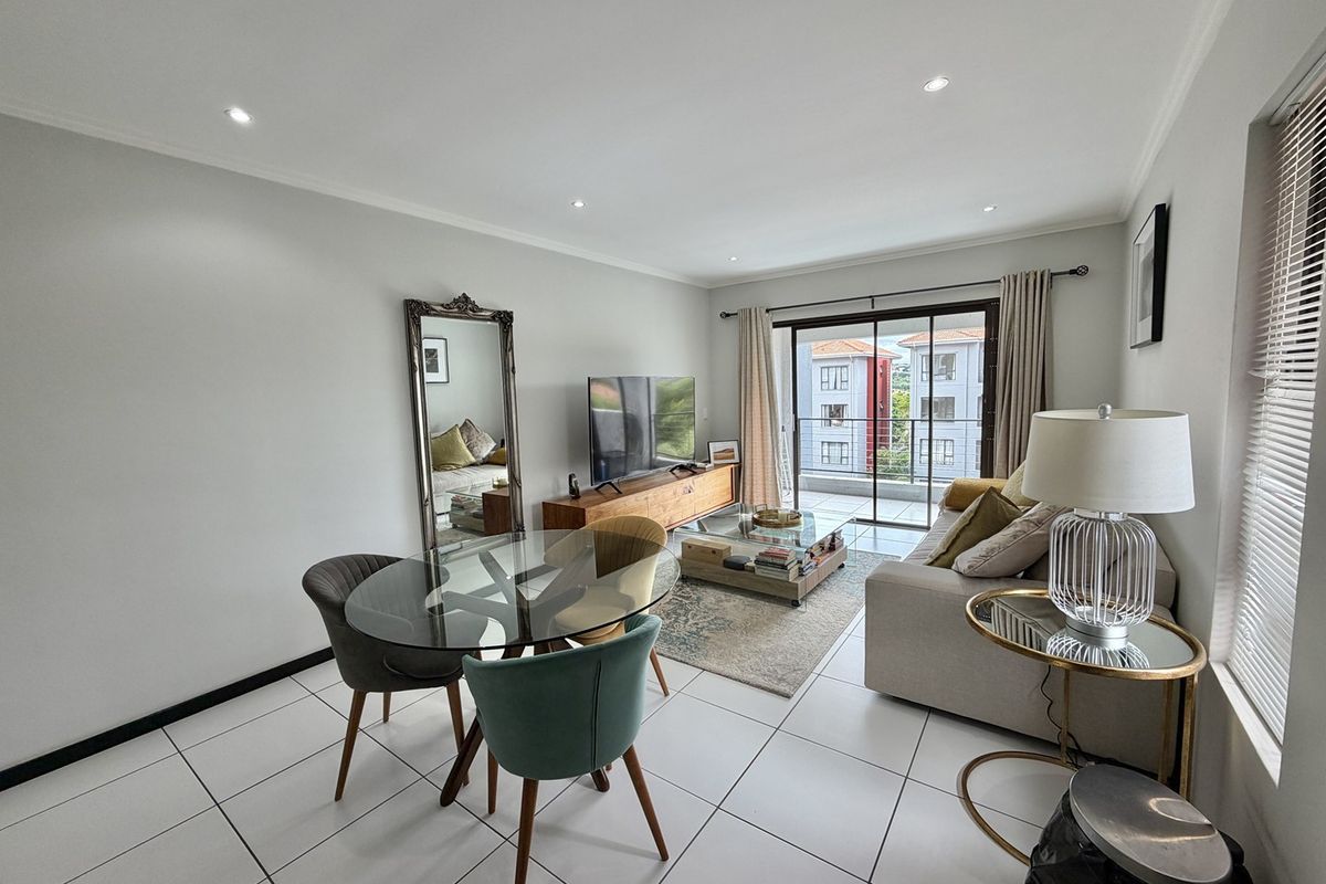 A bright open-plan living and dining area featuring tiled flooring, recessed downlighting, and a sliding glass door leading to a private balcony.