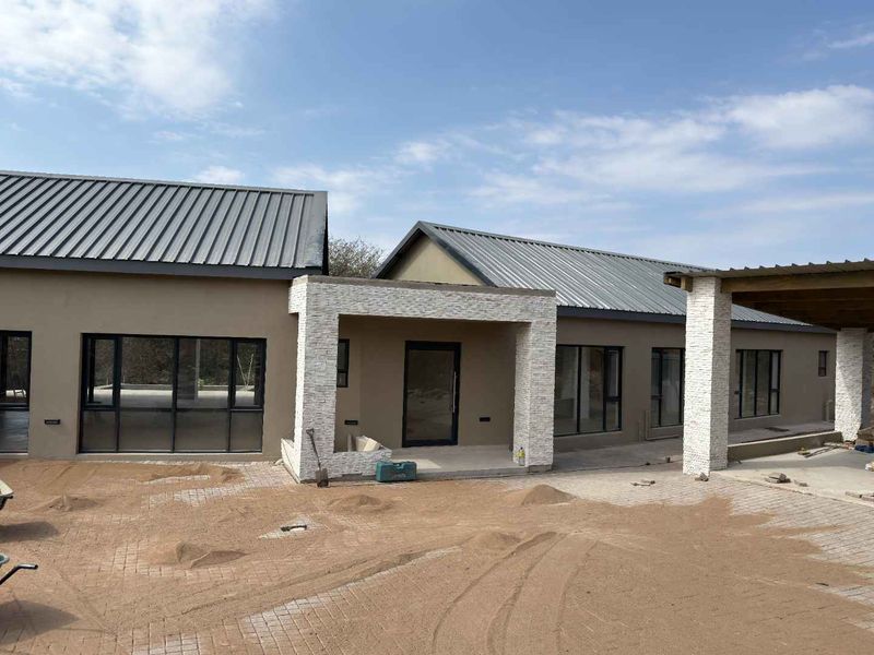 Modern single-story house exterior featuring metal roofing, stone cladding accents, and large aluminium windows, currently undergoing final landscaping and exterior finishing.