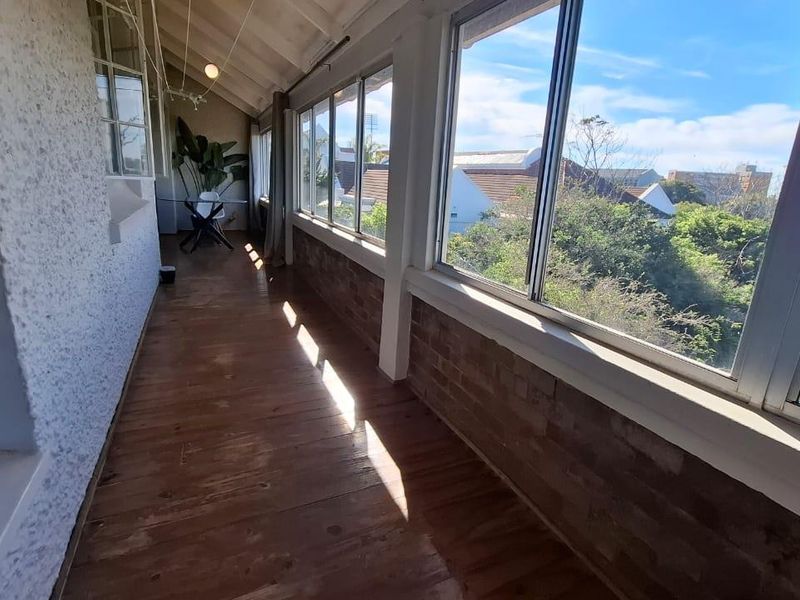 A sunlit enclosed balcony featuring timber flooring, exposed rafters, and expansive windows offering pleasant neighborhood views.