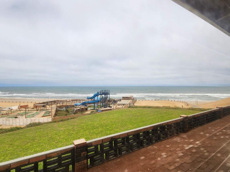 Elevated view overlooking the coastline, beach, and adjacent recreational area, featuring a paved patio with a decorative brick balustrade.
