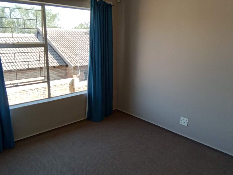 A bright, empty bedroom featuring neutral walls, carpeted flooring, and a large window with security bars, offering a blank canvas for personalization.