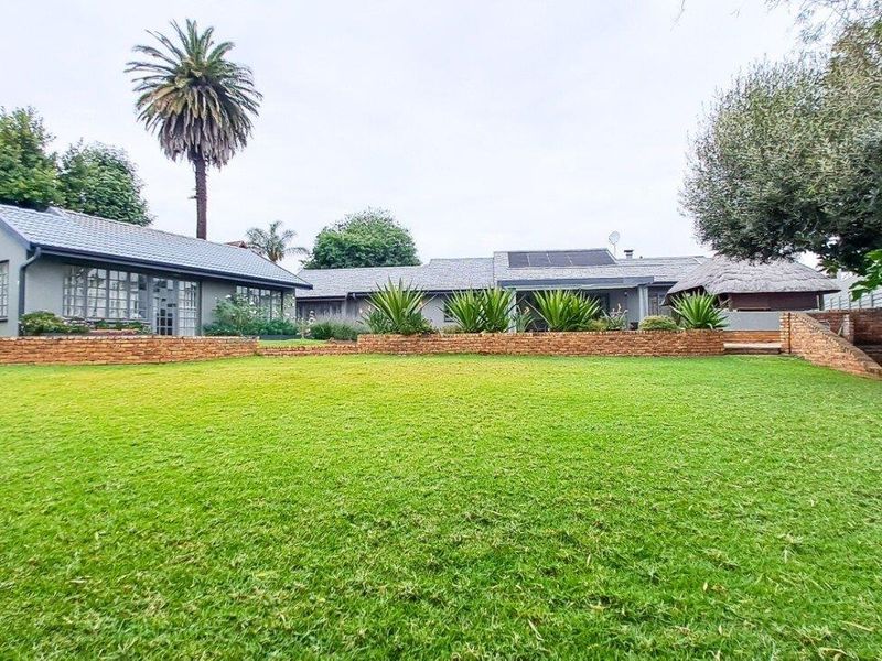 Spacious rear garden featuring a well-maintained lawn, brick retaining walls, and a view of the main residence with solar panels and a thatched roof area.