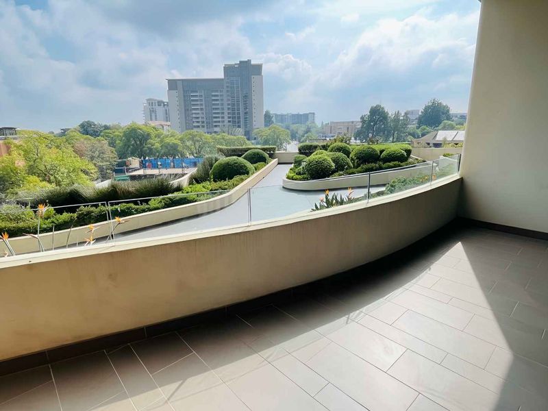 A spacious, modern balcony featuring tiled flooring and a sleek glass balustrade, offering an elevated view of the landscaped gardens and surrounding urban area.