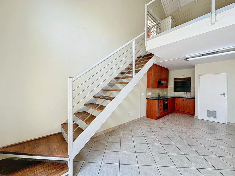 Open-plan living area featuring a mezzanine level, staircase with stainless steel balustrade, and a compact kitchen with built-in cabinetry and appliances.