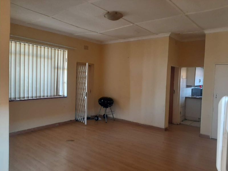 Spacious open-plan living area featuring laminate flooring, vertical blinds, and a security gate, with an adjacent kitchen visible in the background.