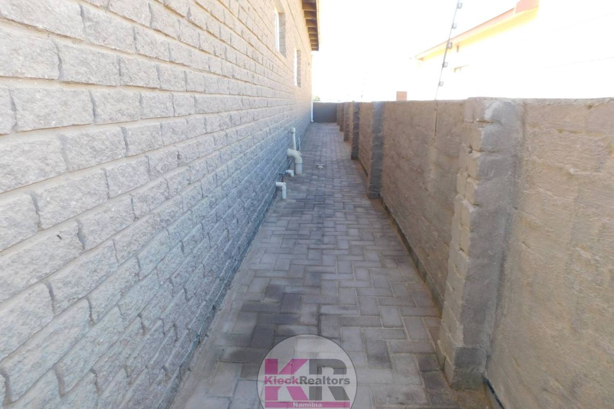A narrow paved side passage featuring brick boundary walls, exposed plumbing infrastructure, and security electric fencing.