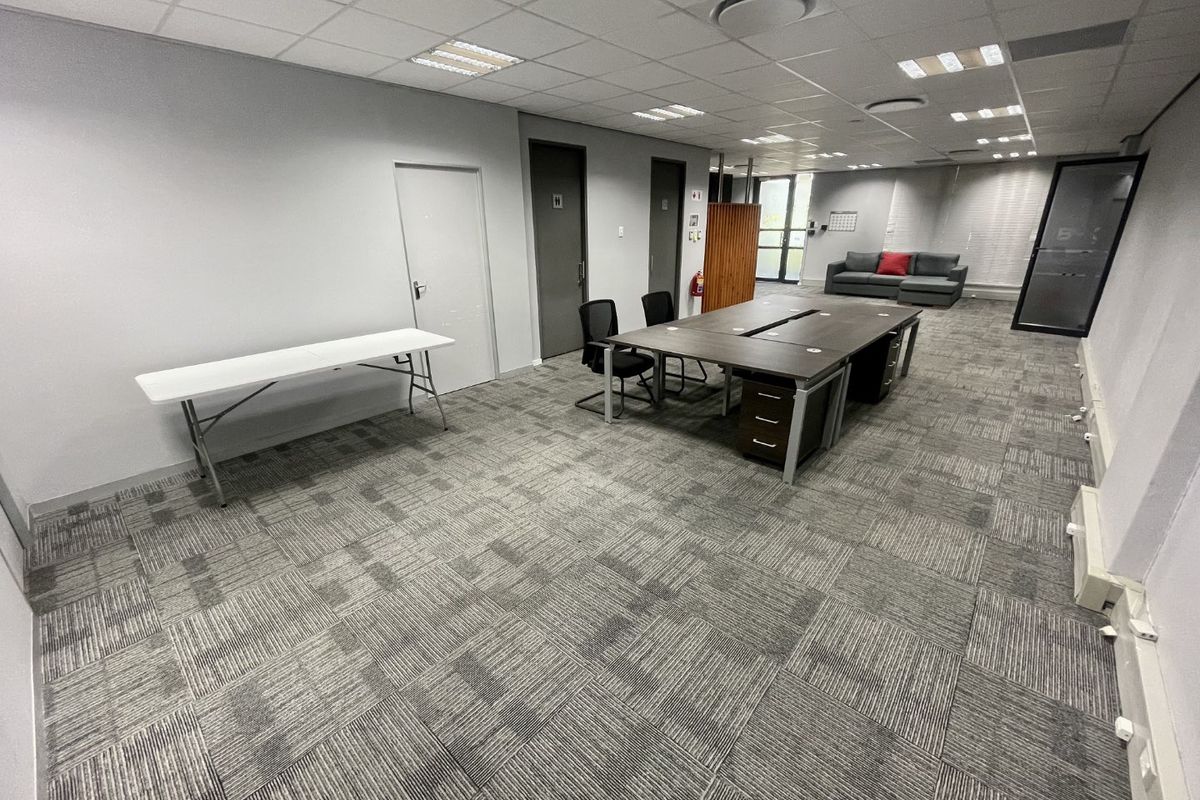 A spacious open-plan office area featuring carpet tile flooring, suspended ceiling with recessed lighting, and glass partition elements.