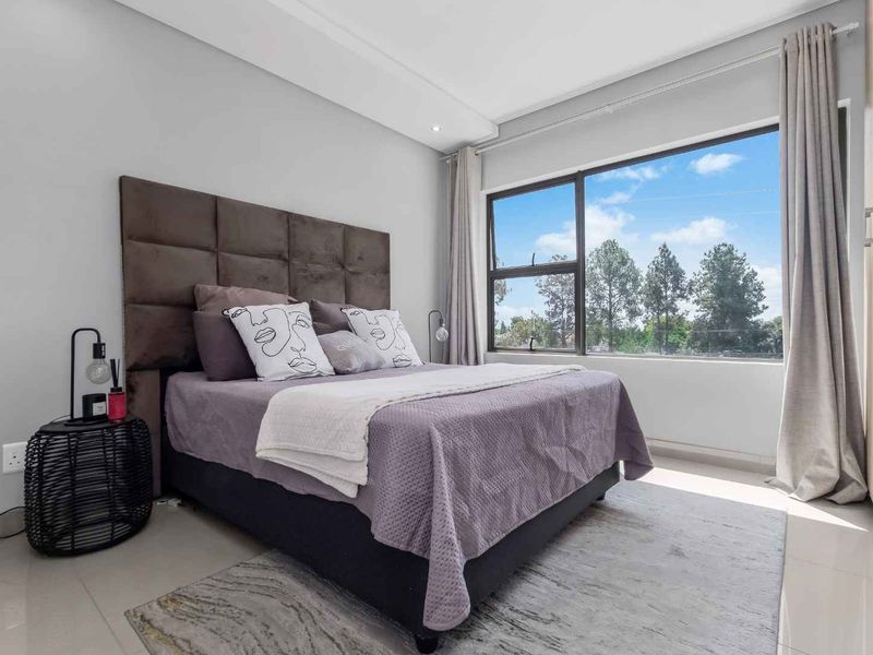 A bright and modern bedroom featuring tiled flooring, built-in cupboards, and a large window providing natural light and pleasant outdoor views.