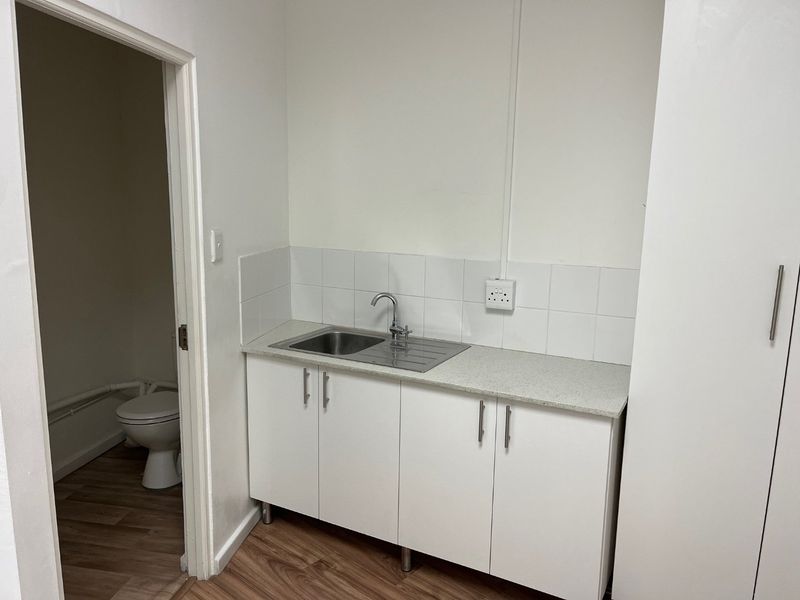 A compact kitchenette featuring white cabinetry, a stainless steel sink, and a tiled backsplash, with an adjacent small bathroom visible through the doorway.