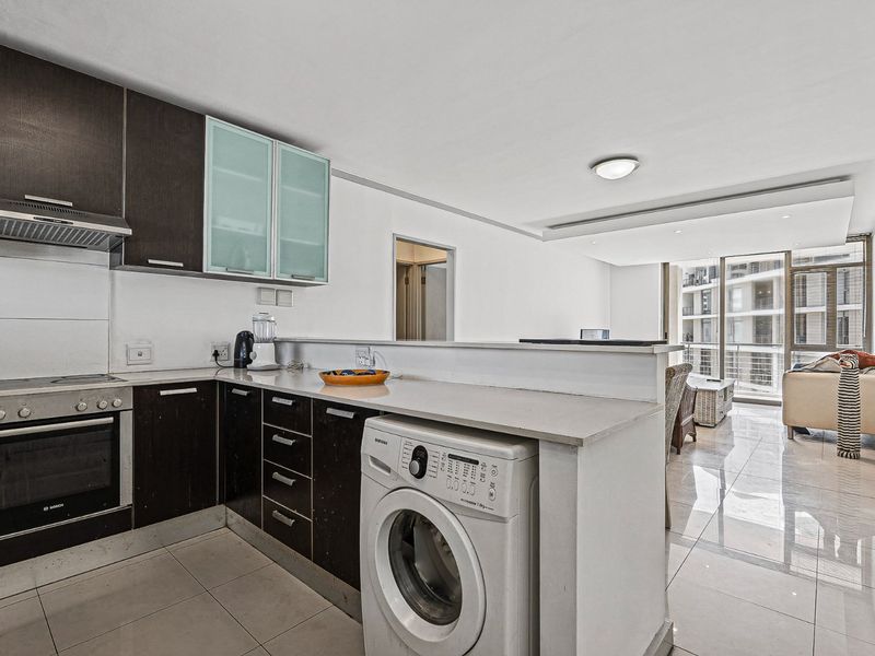 Modern open-plan kitchen featuring dark cabinetry, stone countertops, and integrated appliances, flowing seamlessly into a bright living area with large sliding glass doors.