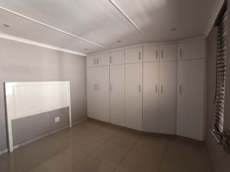 A clean, empty bedroom featuring extensive white built-in cupboards, neutral tiled flooring, and modern recessed lighting.