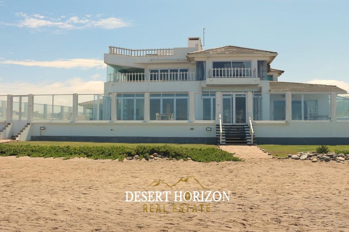 A beachfront guest house featuring a multi-level exterior with glass balustrades, expansive windows, and direct access to the sandy beach.